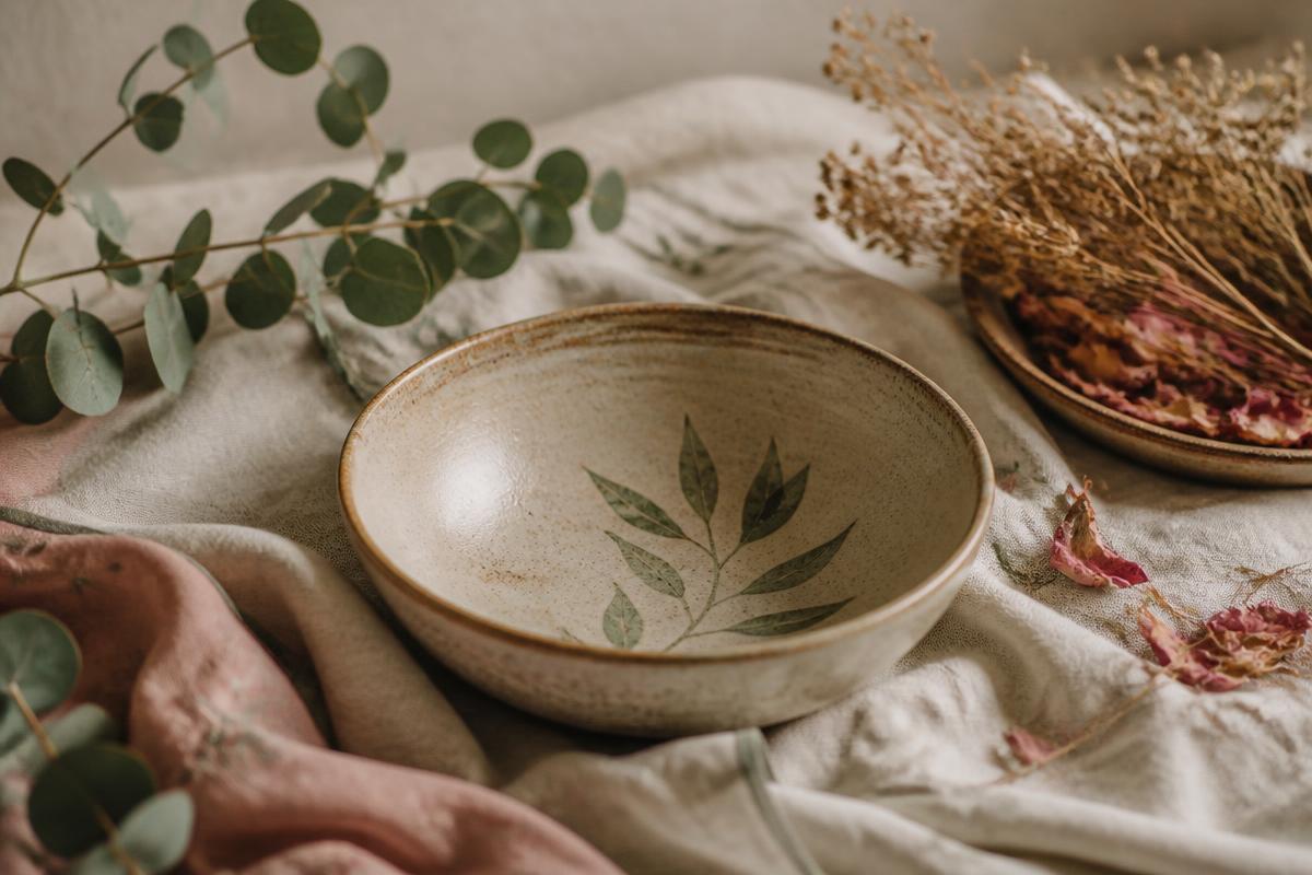 A handmade ceramic bowl resting on linen with eucalyptus leaves and dried rose petals