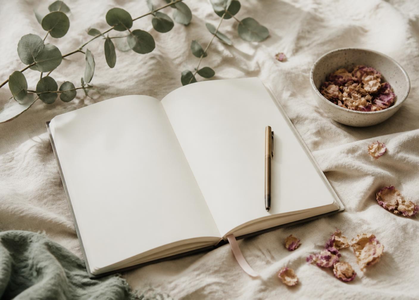 An open journal with a pen, dried rose petals and eucalyptus on cream linen